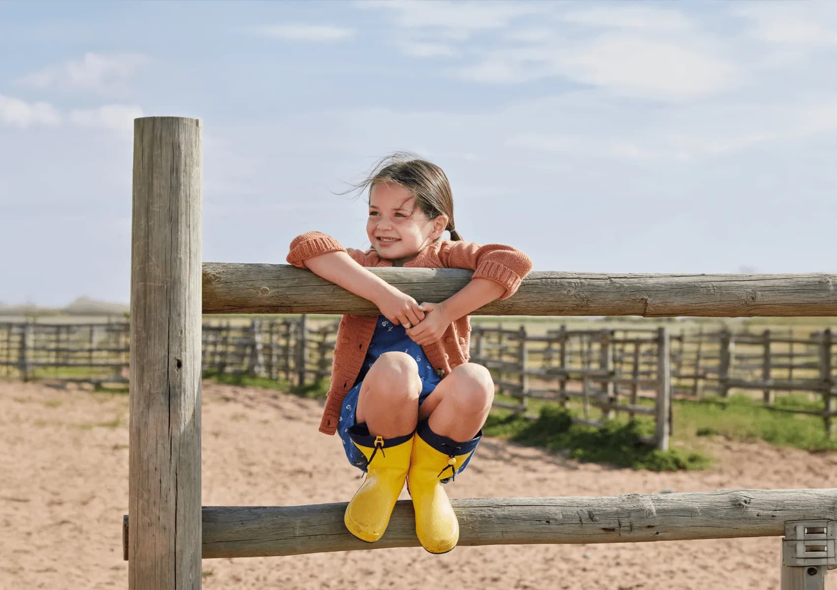 A child in yellow boots sitting on a farm fence.